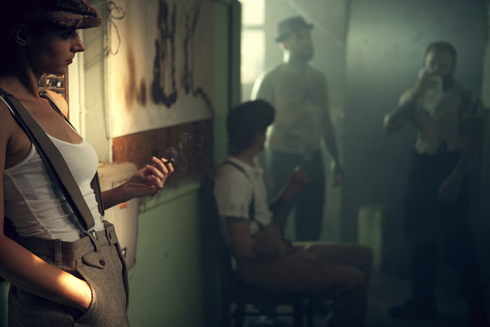 A woman in a flat cap smokes in the foreground while the men wait behind