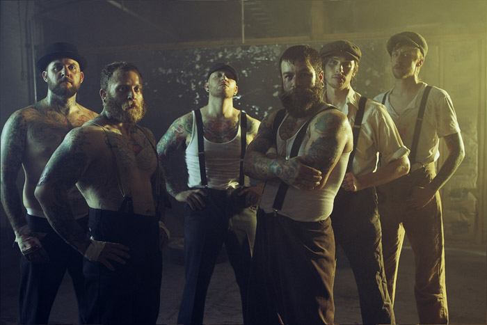 Six men in suspenders and flat caps, tattooed, in a warm-lit warehouse
