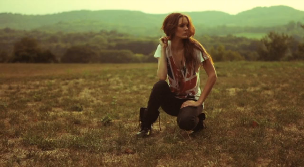 The model kneeling in a field with hills in the distance